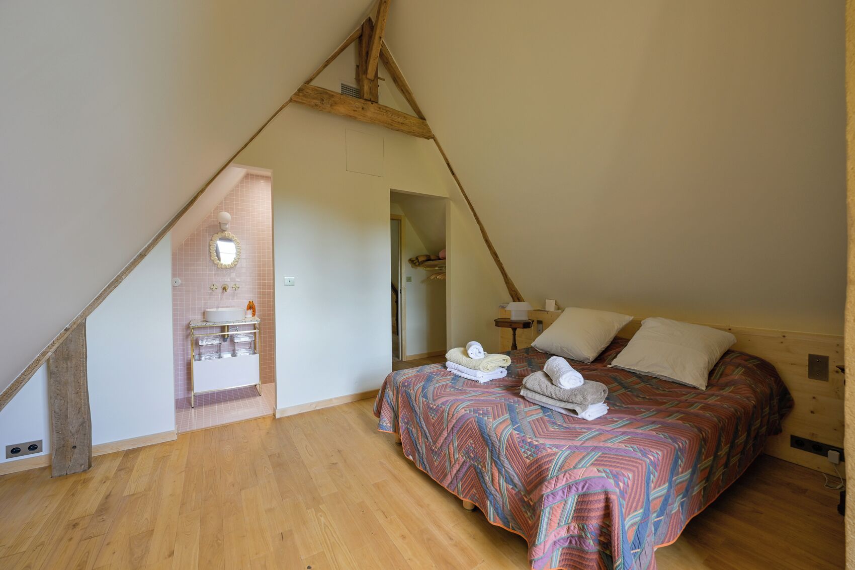 A bedroom with a sloped ceiling, a bed with a patterned bedspread, and a small bathroom with a sink and mirror.