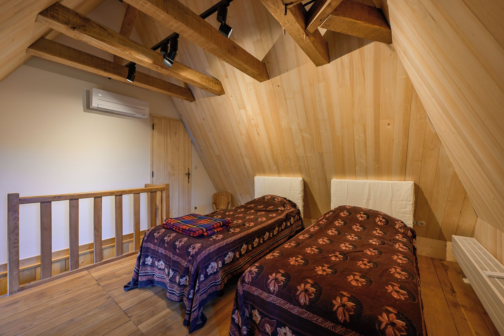 Une petite chambre mansardée en bois, avec deux lits, une rambarde en bois et un climatiseur blanc.