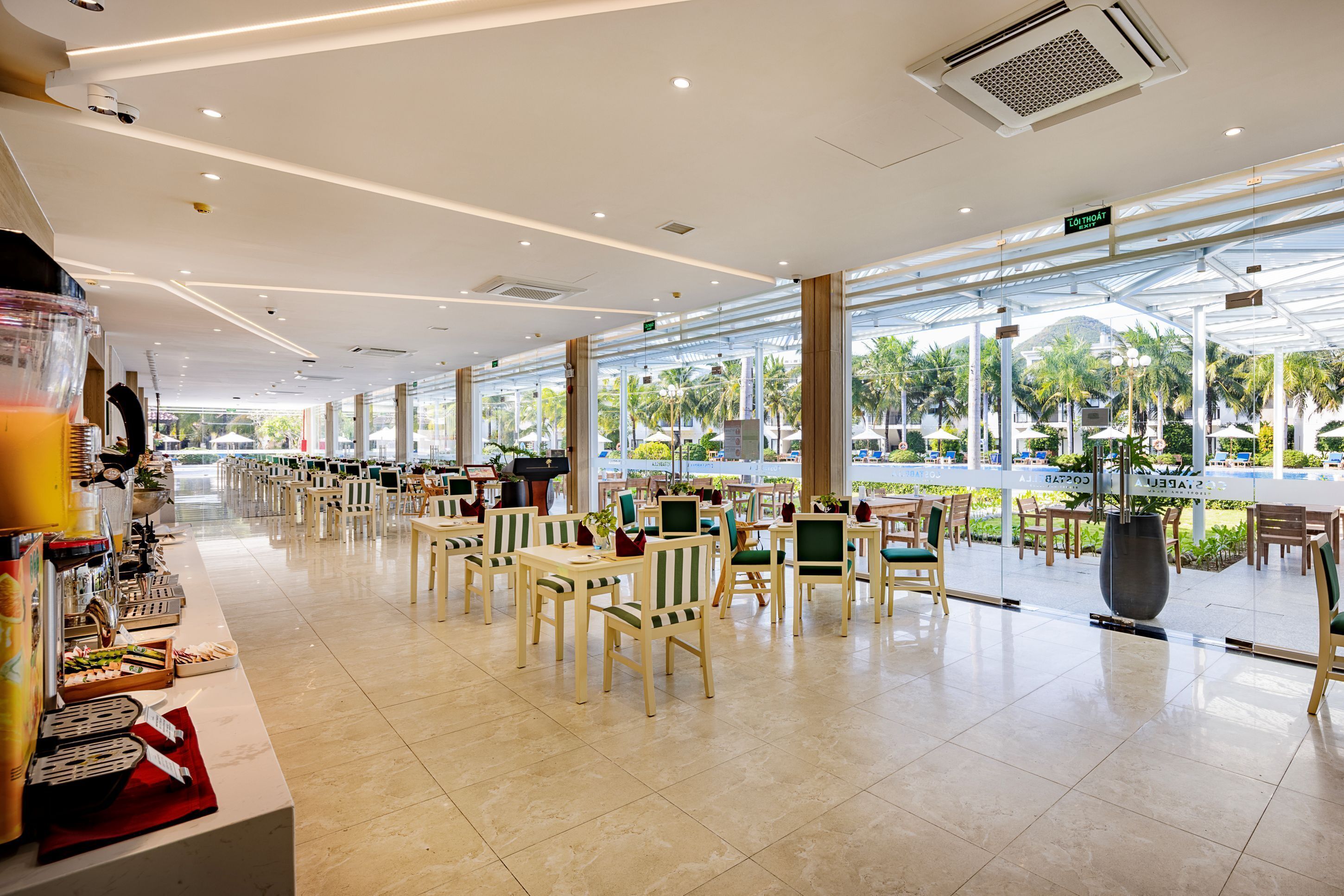A spacious indoor area with multiple tables and chairs, large windows, and a view of palm trees and a pool outside.