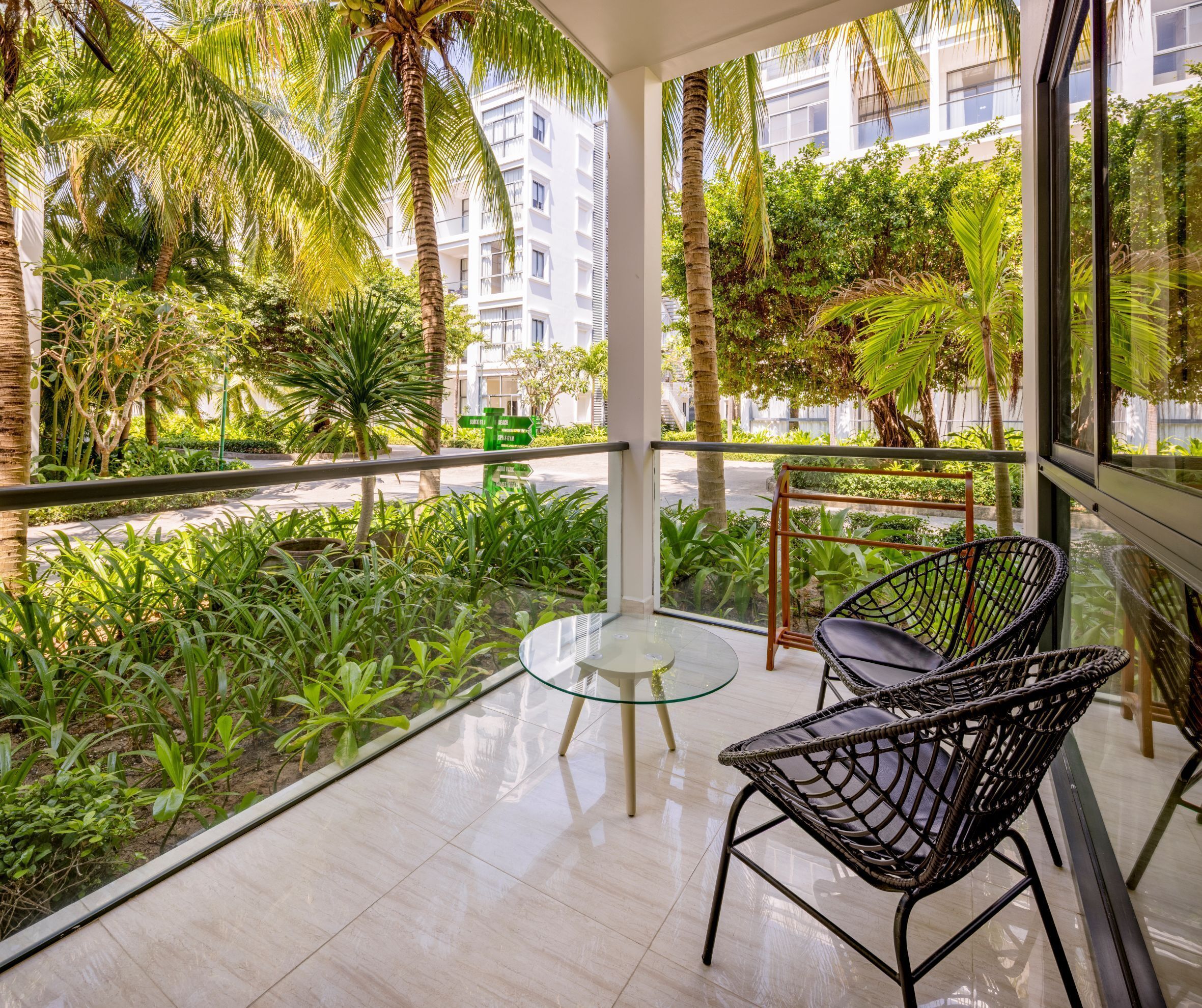 A balcony with a glass table, two wicker chairs, and lush greenery.