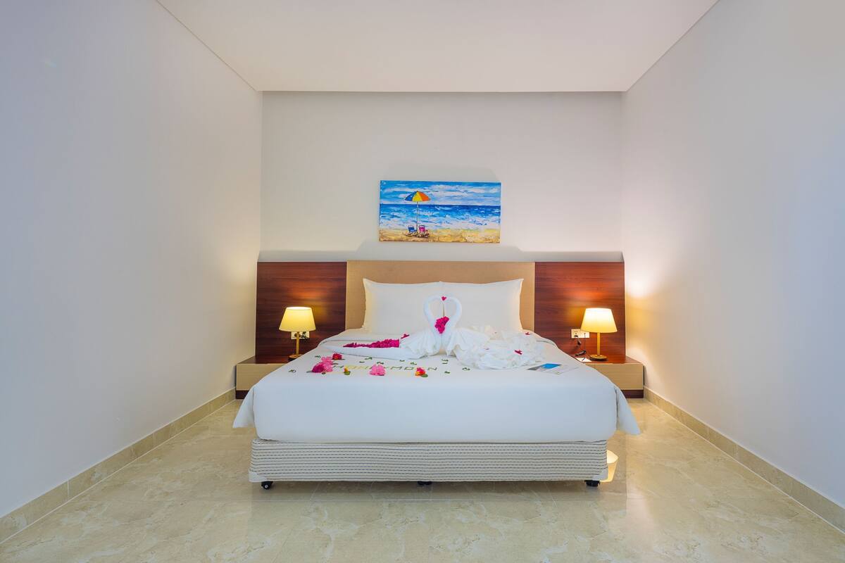 A hotel room with a bed, two bedside lamps, a painting above the bed, and a small table with a lamp.