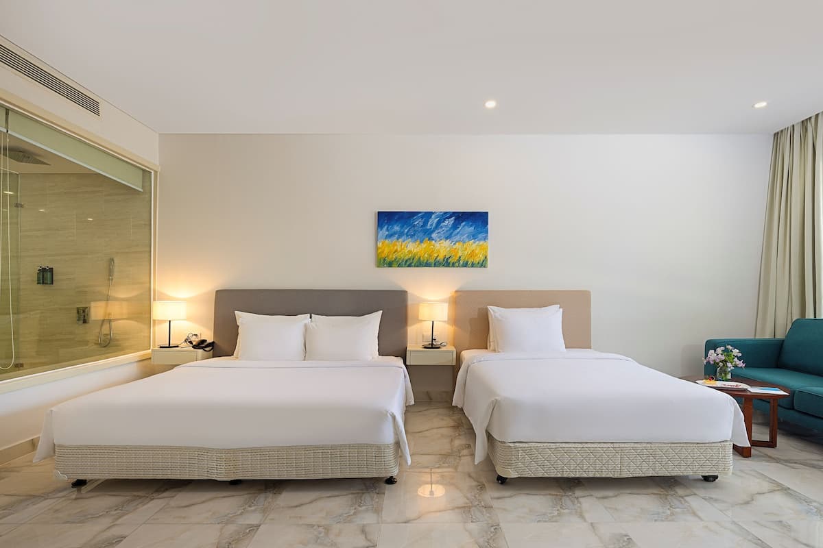 A hotel room with two beds, a glass shower enclosure, a painting on the wall, and a green armchair.