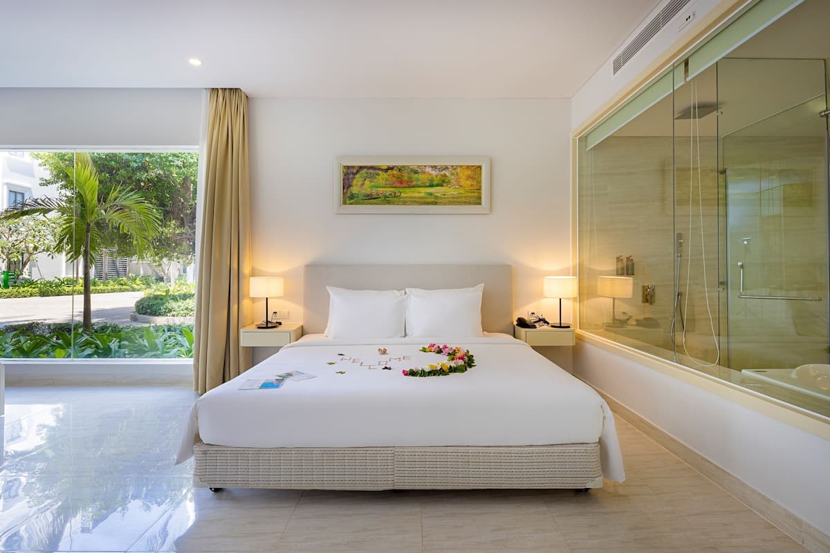 A hotel room with a large bed, a glass shower enclosure, and a view of greenery outside.