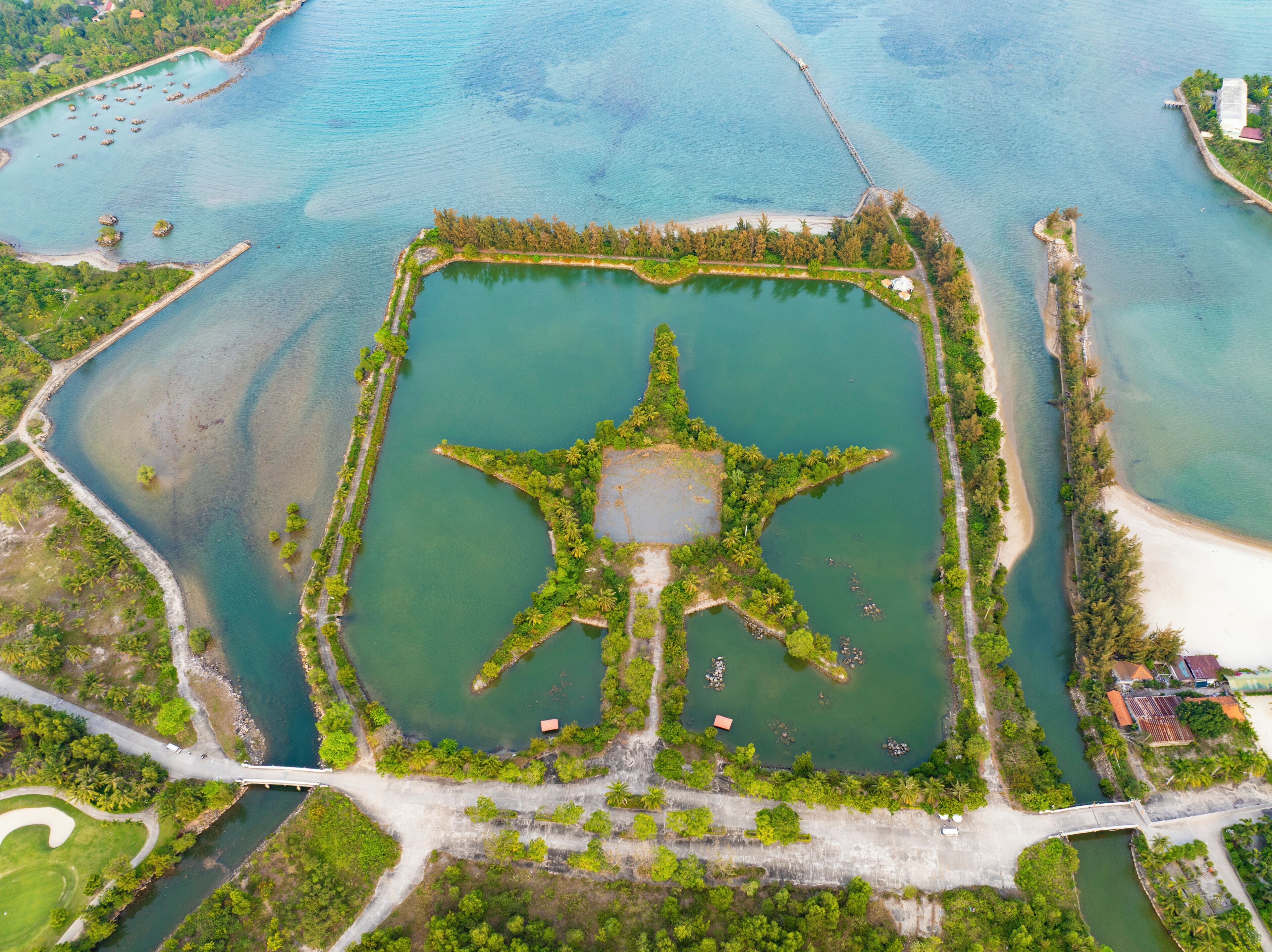 Eine Luftaufnahme eines quadratischen Wasserbeckens mit einer sternförmigen Insel in der Mitte, umgeben von Grünflächen und verbunden durch Brücken.