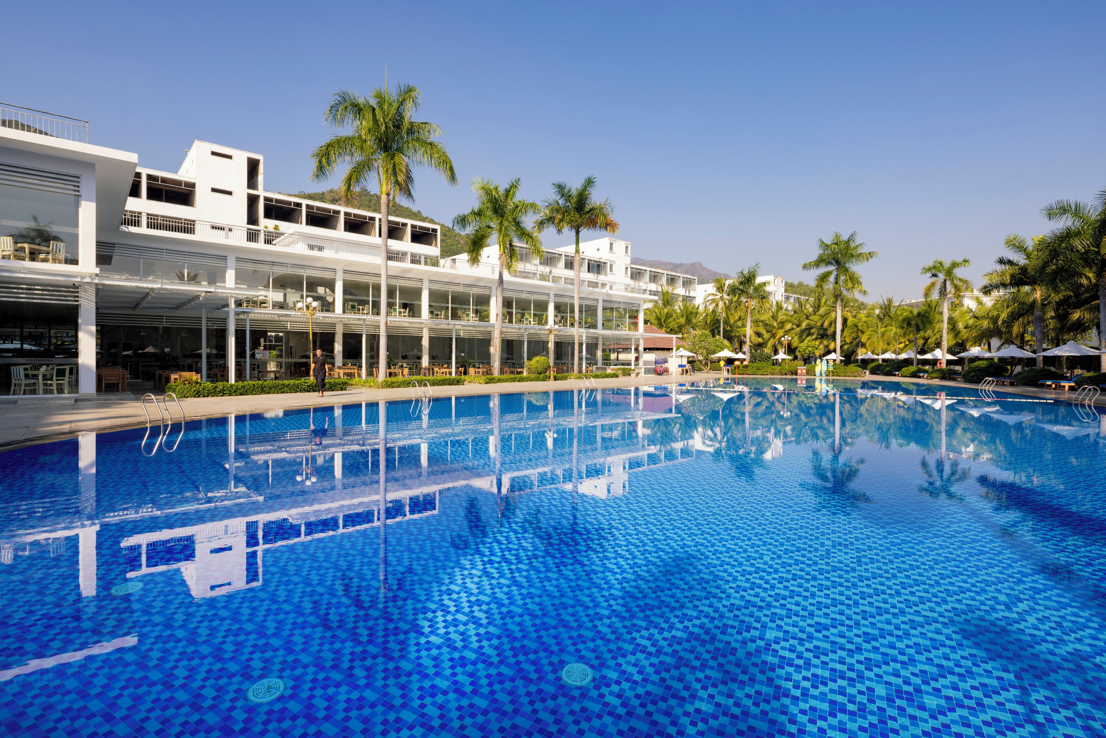 A swimming pool with a tiled floor, surrounded by a building with multiple balconies and palm trees.