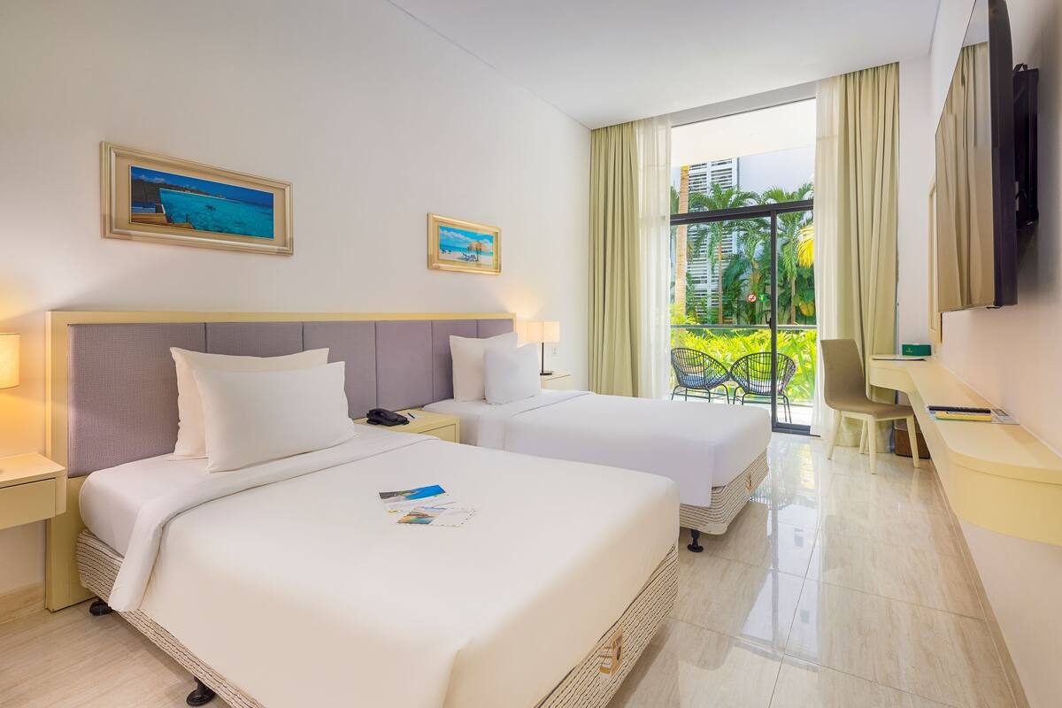 A hotel room with two beds, a desk, a chair, a TV, and a balcony with a view of buildings and greenery.