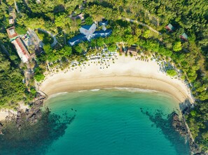 An aerial view of a beach with a curved shoreline, surrounded by dense greenery and a few buildings.