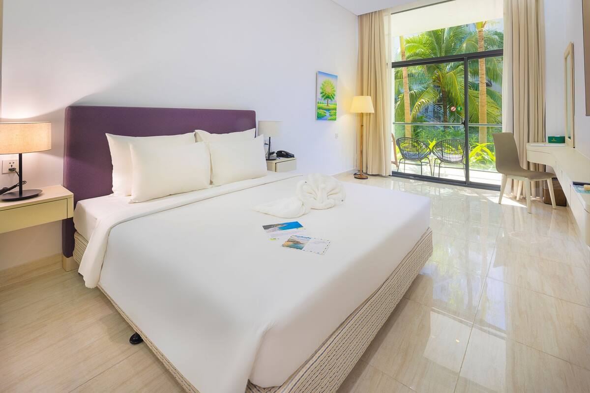 A hotel room with a large bed, a nightstand, a desk, and a view of palm trees through the window.