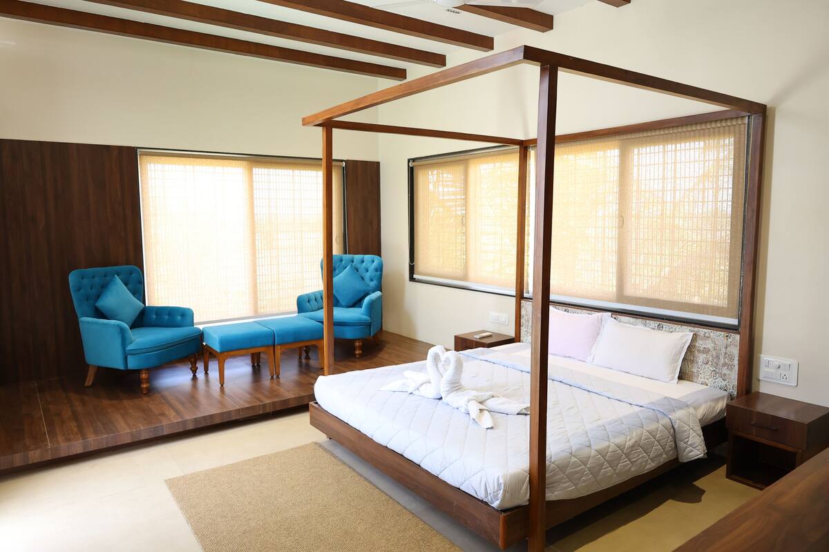 Une chambre à coucher avec un lit à baldaquin, deux fauteuils bleus, un plancher en bois et de grandes fenêtres équipées de stores.