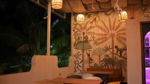 A restaurant with wooden tables and chairs, a mural with tropical wildlife, and hanging lights. - Coconut Lagoon Agro Resort (Parbhani)