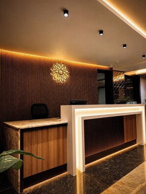 A modern reception desk with a wooden panel design, a logo on the wall, and a reception area with a computer and a plant. - Sainik Grand (Varanasi)