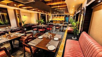 A restaurant with multiple dining tables, red cushioned seating, and a kitchen area in the background.