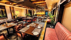 A restaurant with multiple dining tables, red cushioned seating, and a kitchen area in the background. - Sainik Grand (Varanasi)