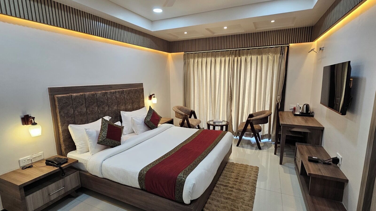 A modern hotel room with a large bed, bedside tables, a desk, a chair, a television, and a window with curtains.