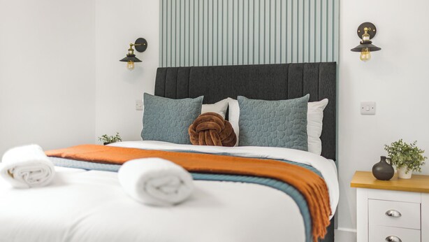 Un letto rifatto con cura, con biancheria da letto bianco e arancione, una testata scura e due lampade a parete.