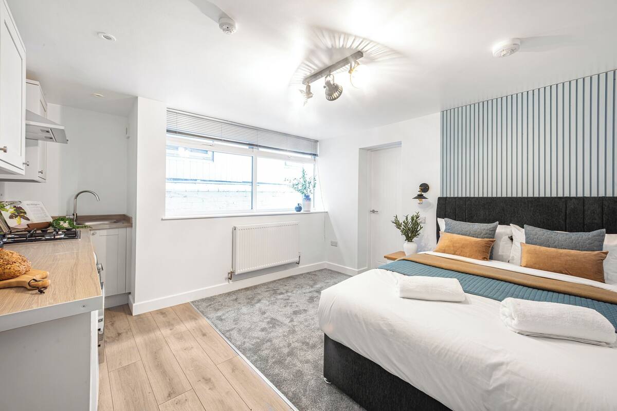 A modern bedroom with a large bed, a built-in desk, a window with blinds, and a radiator.