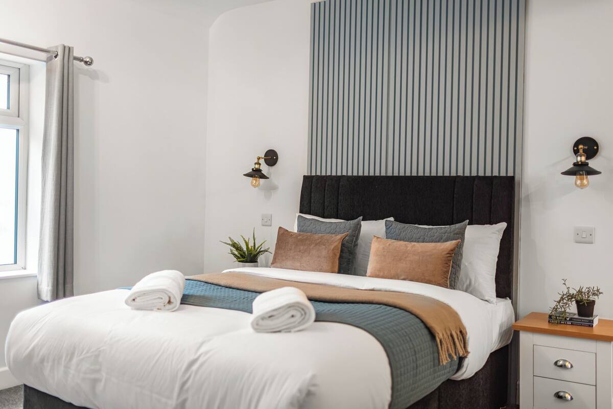 A modern hotel room with a large bed, a nightstand, a potted plant, and a window with curtains.