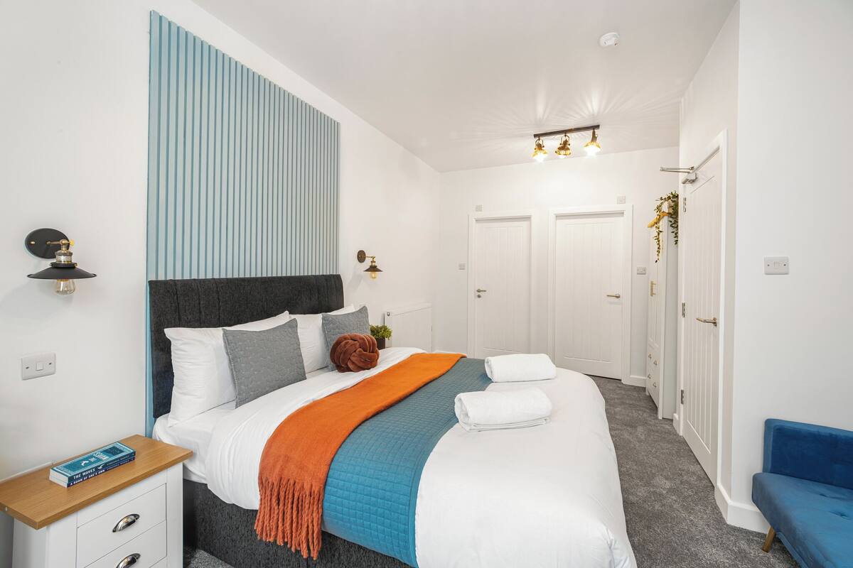 A neatly arranged hotel room with a large bed, a bedside table with a book, a blue chair, and a wardrobe.