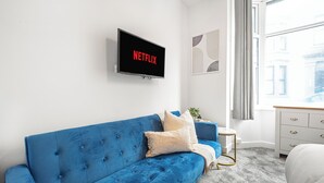 Una camera da letto moderna con un divano blu, una TV a parete che mostra 'NETFLIX', un piccolo tavolino con una pianta e una finestra con vista sugli edifici.