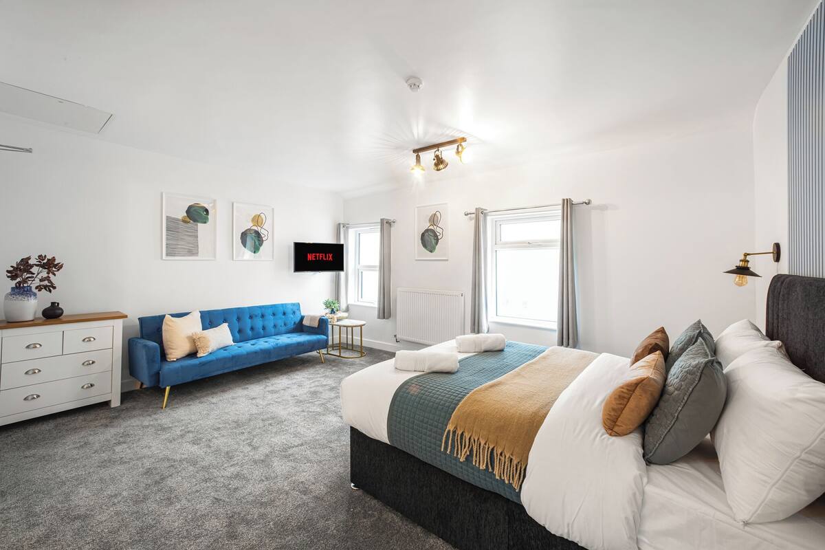 A modern bedroom with a large bed, a blue sofa, a white dresser, and a flat-screen TV mounted on the wall.