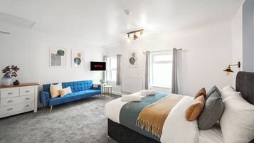 A modern bedroom with a large bed, a blue sofa, a white dresser, and a flat-screen TV mounted on the wall.
