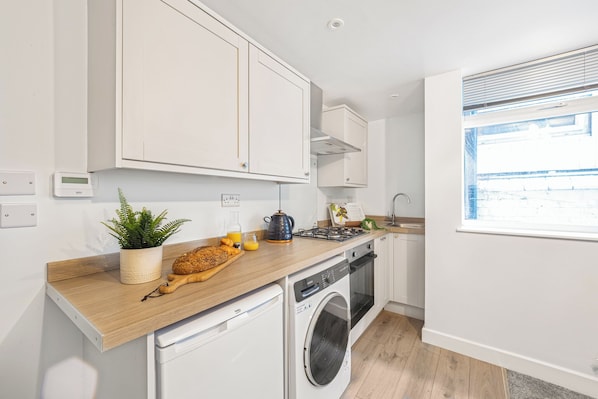 A modern kitchen with white cabinets, a wooden countertop, a washing machine, and a window with blinds. - Aurum House (Blackpool)