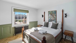 A bedroom with a large bed, a radiator, a window with a green valance, a painting on the wall, and a nightstand with a lamp. - Aldwark Mews (York)
