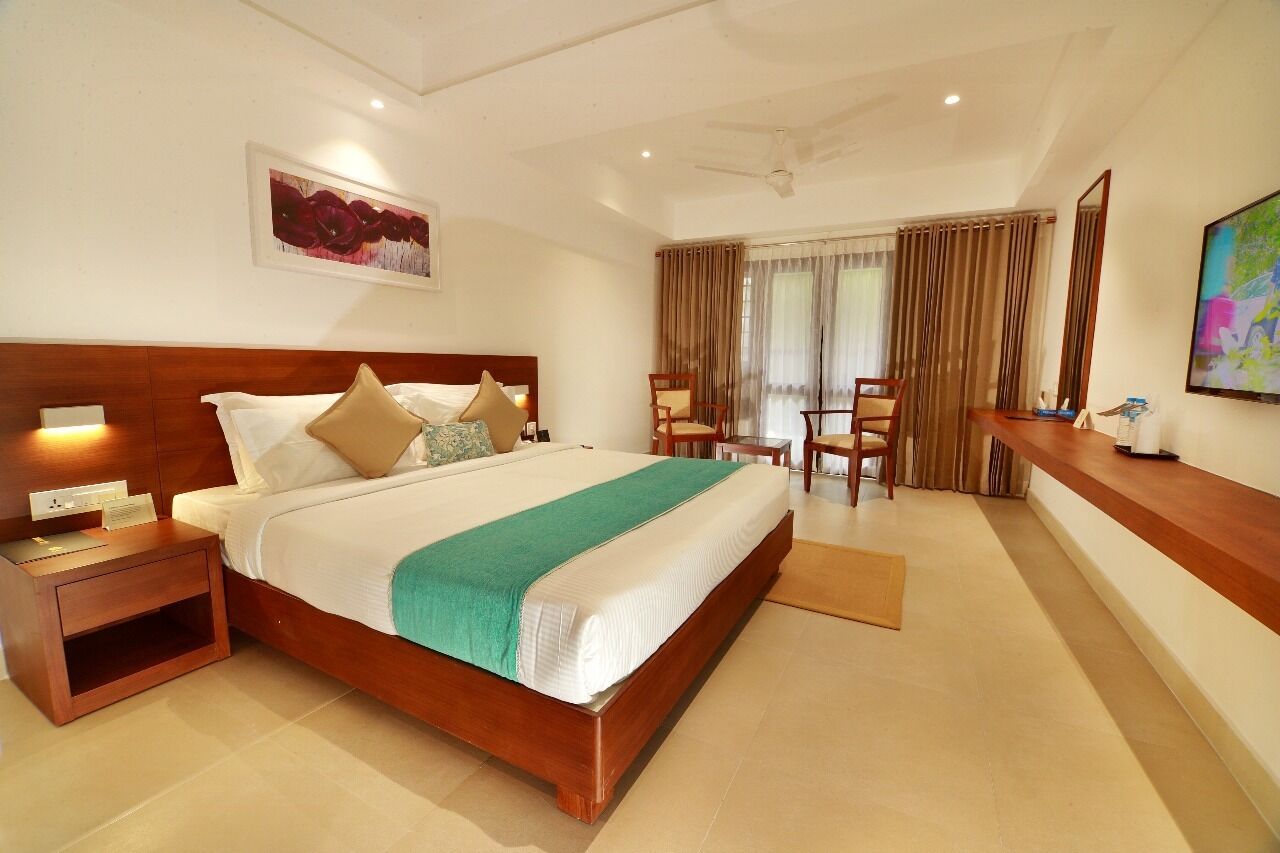 A hotel room with a large bed, a wooden nightstand, a desk with a chair, and a large window with curtains.