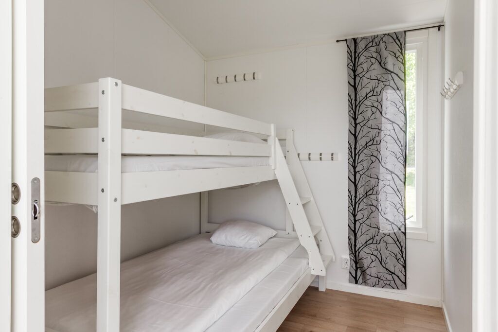 Ein Schlafraum mit Etagenbetten, weißen Wänden, einem Fenster mit Vorhang mit Baumdekor und Holzboden.