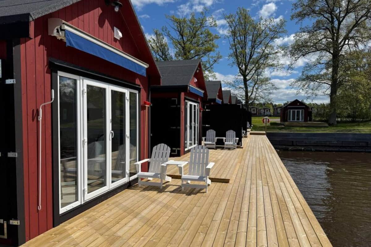 Une terrasse en bois avec des chaises blanches et une table, qui mène à un bâtiment rouge doté de portes vitrées.
