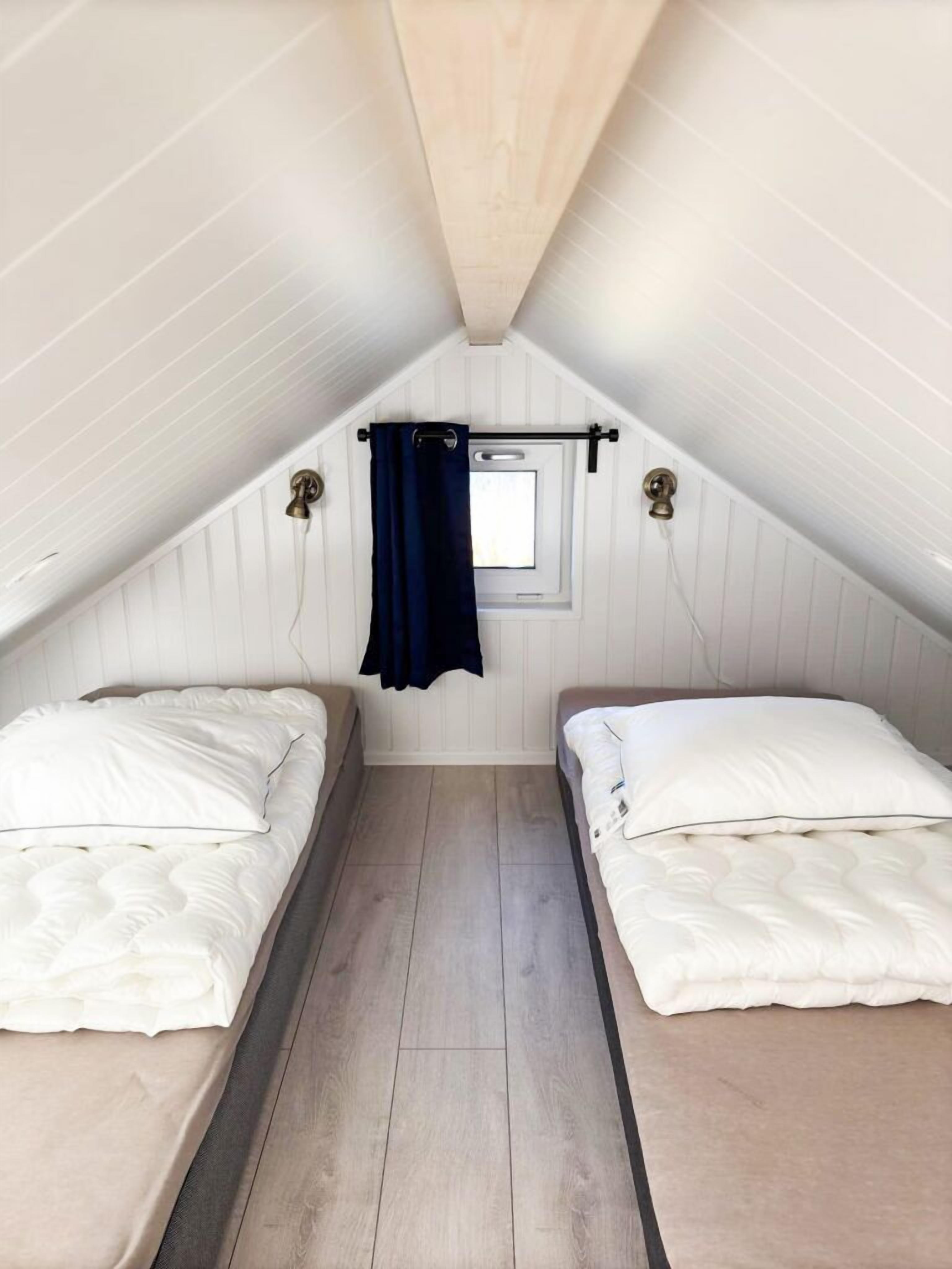 A small attic bedroom with two beds, wooden flooring, and a window with curtains.