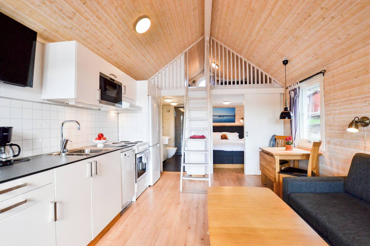 A compact, modern kitchen with white cabinets, a microwave, and a stove. There is a wooden dining table with a chair and a sofa in the living area. A staircase leads to an upper level with a bed and a window.