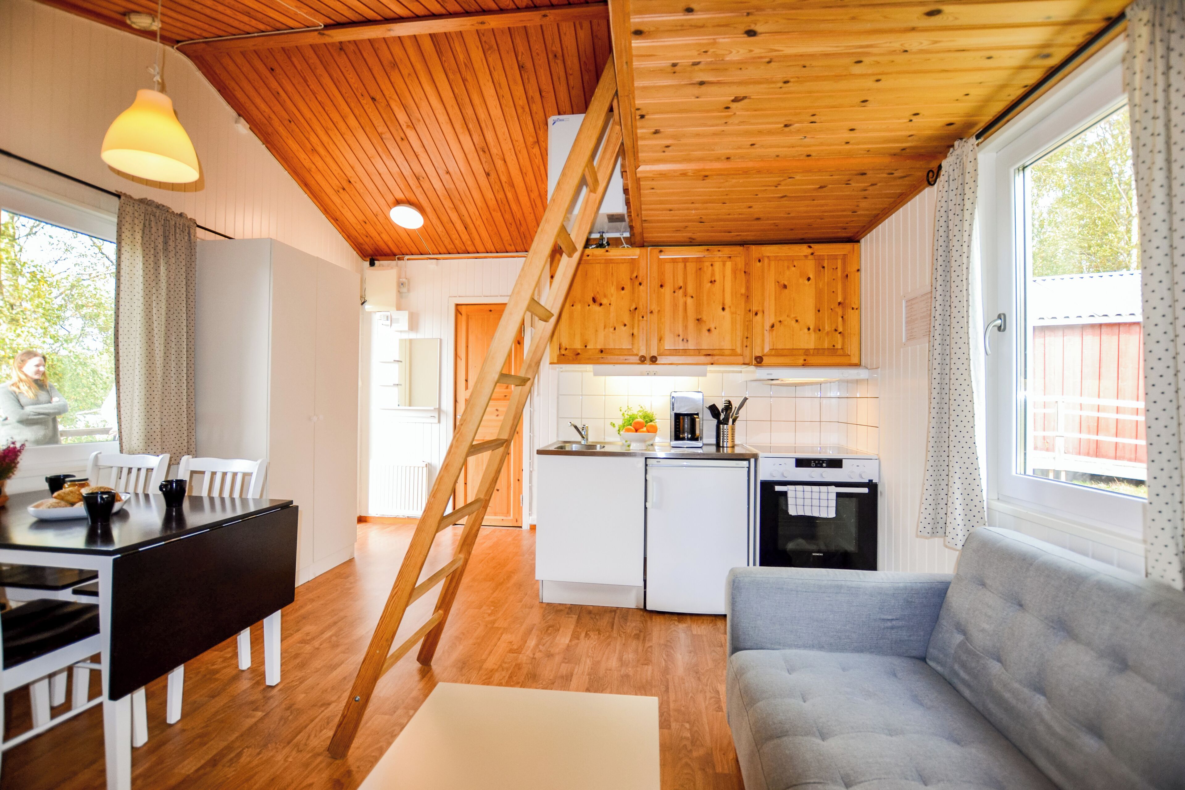 A modern kitchen with wooden cabinets, a white refrigerator, and a coffee maker. There is a wooden ladder leading to a lofted area.
