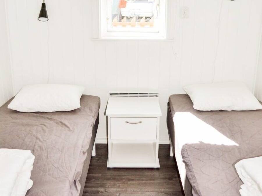 Une chambre avec deux lits, un petit bureau blanc, une fenêtre avec un store noir et une lampe fixée au mur.
