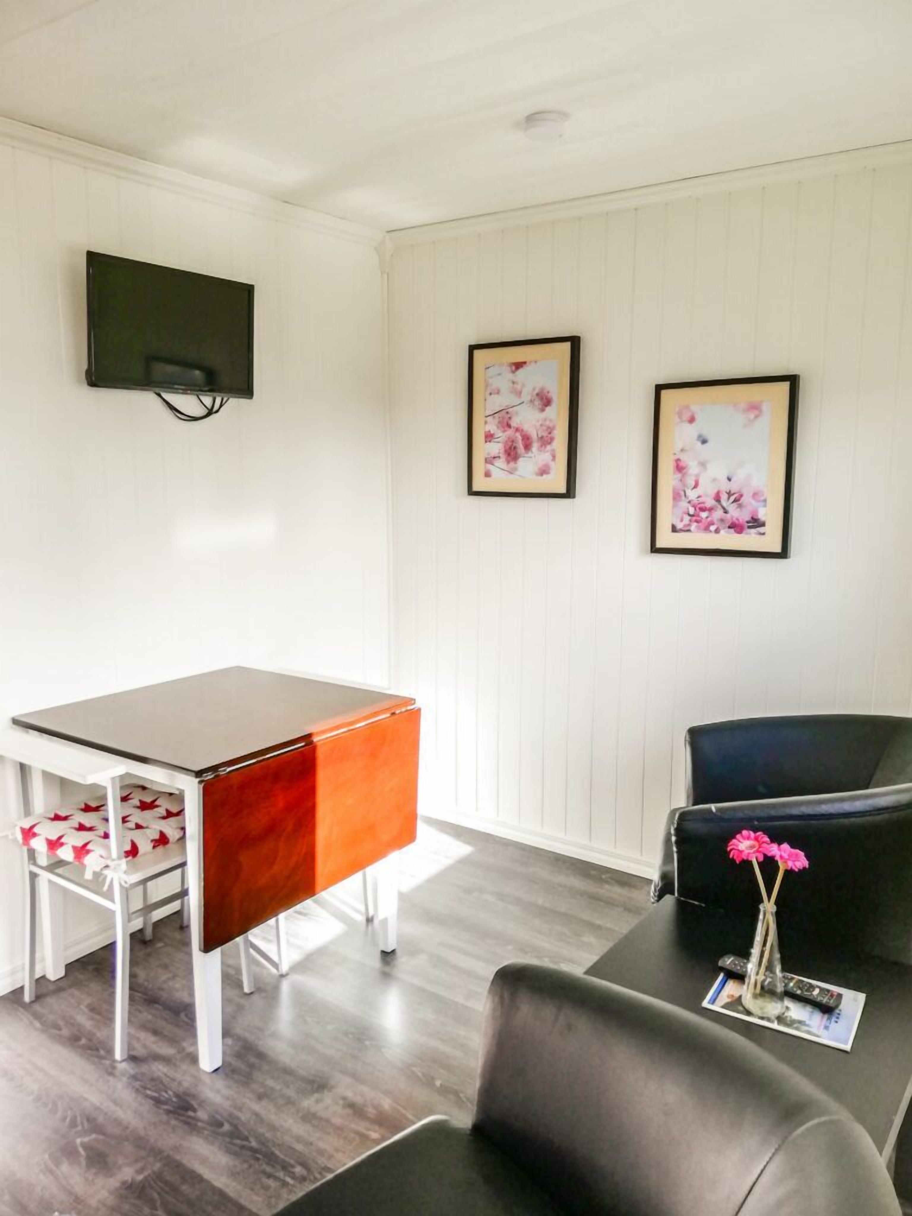 Ett rum med en vit vägg, en platt-TV som hänger ovanför ett skrivbord i trä, två inramade konstverk med blommotiv, en svart lädersoffa och ett litet bord med en vas med blommor.