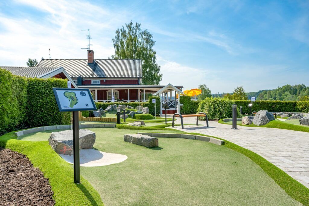 Ett välskött trädgårdsområde med en gångstig, en röd byggnad och en vägskylt.