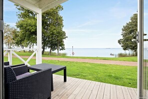 A covered outdoor seating area with wicker furniture, a wooden deck, and a view of a lake with a path and trees. - First Camp Vadstena - Vättern (Vadstena)