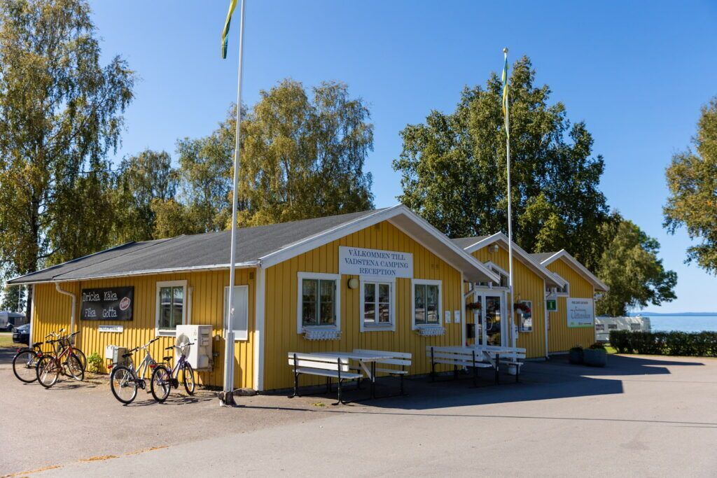 Un edifici groc amb un cartell que diu "Väklommenn till Västena Camping Reception". Hi ha bicicletes aparcades a fora i bancs al davant.