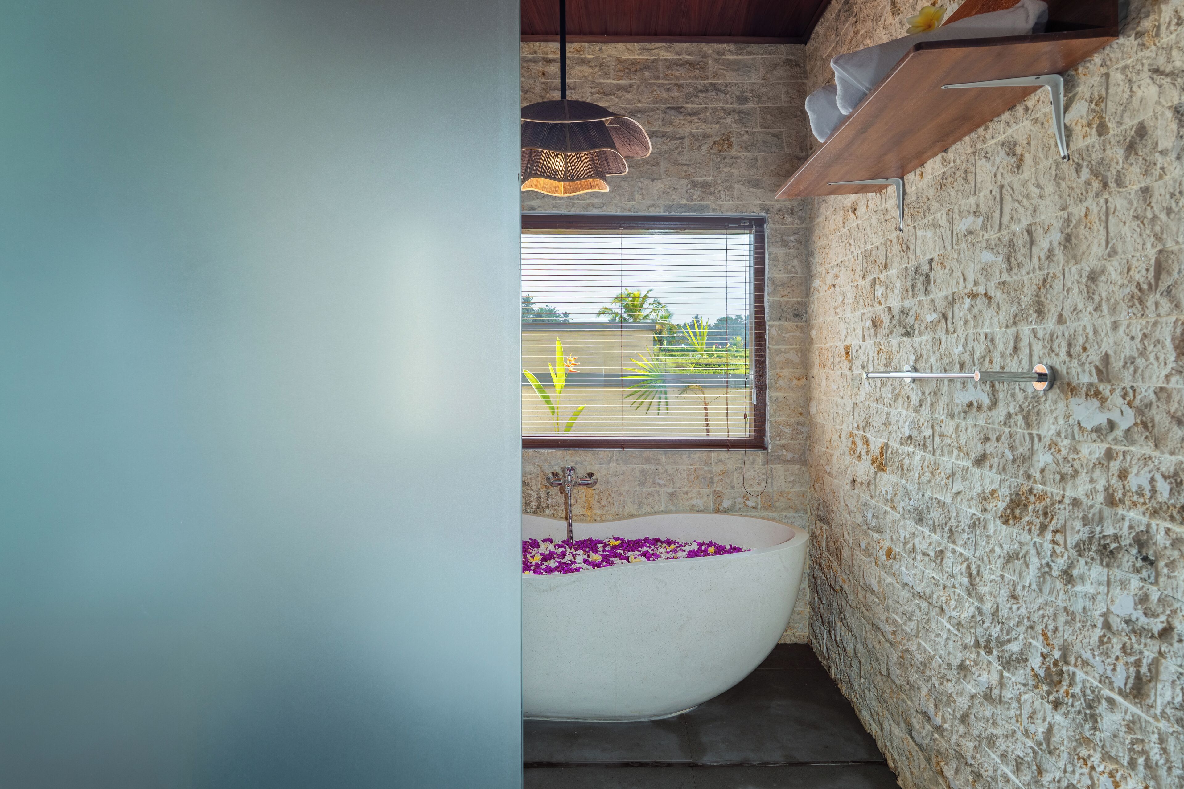 Une salle de bain avec un mur en pierre, une baignoire blanche remplie de fleurs, une fenêtre avec des stores et un luminaire suspendu.