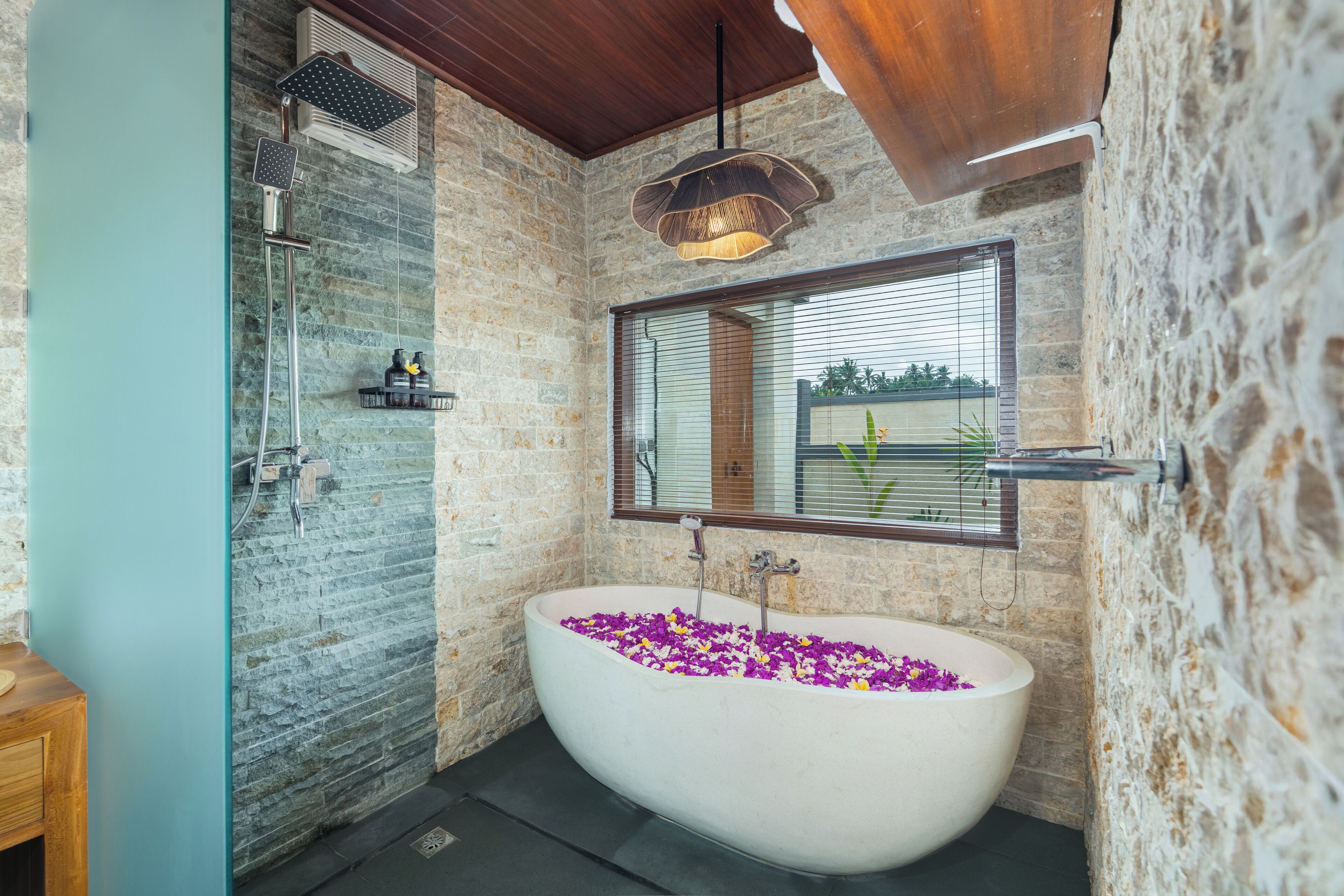 Un baño con una bañera independiente llena de flores moradas, ducha y un amplio ventanal con persianas.
