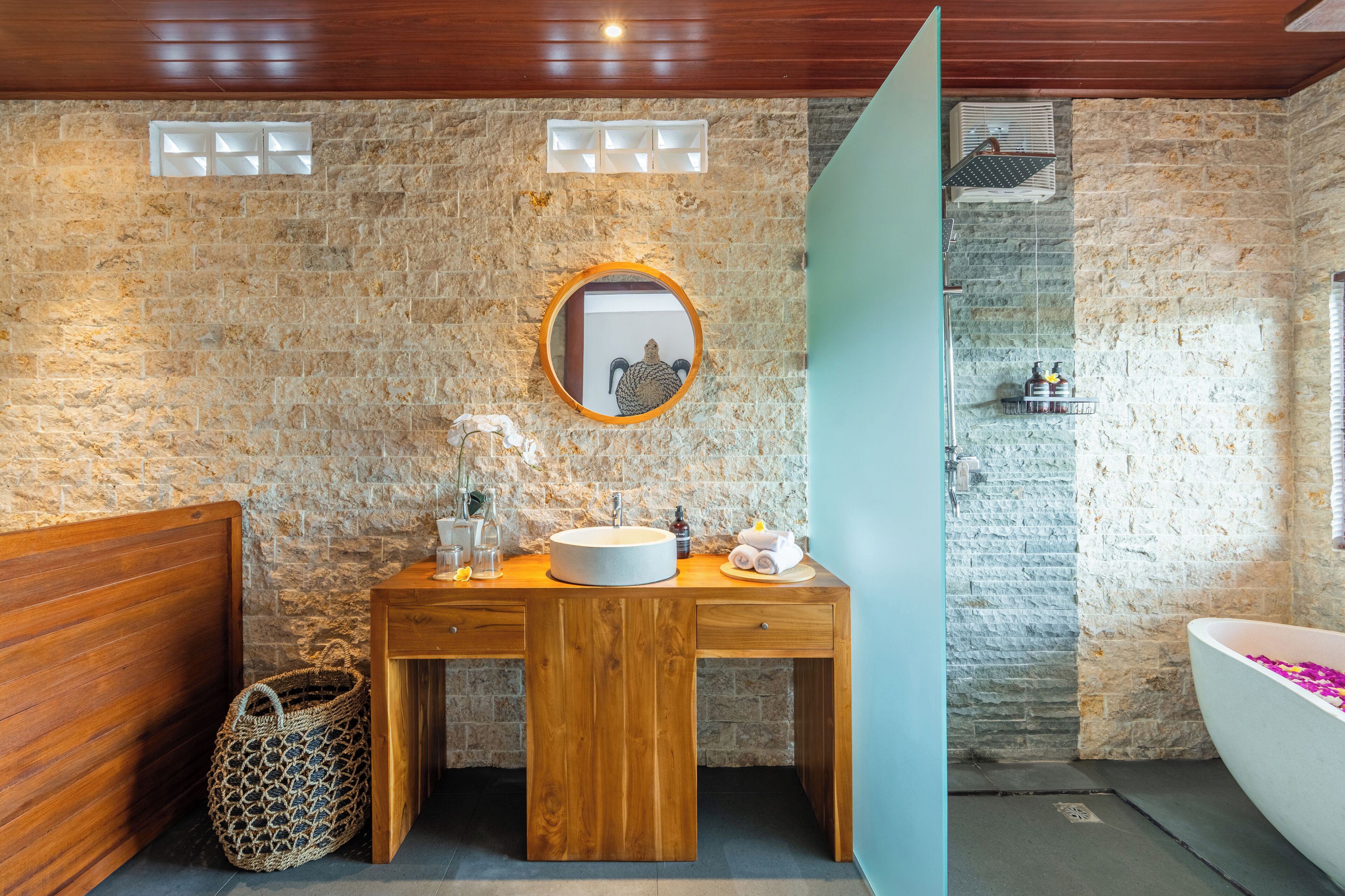 A bathroom with a stone wall, wooden vanity, round mirror, and a freestanding bathtub.