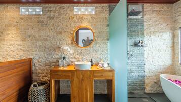A bathroom with a stone wall, wooden vanity, round mirror, and a freestanding bathtub.