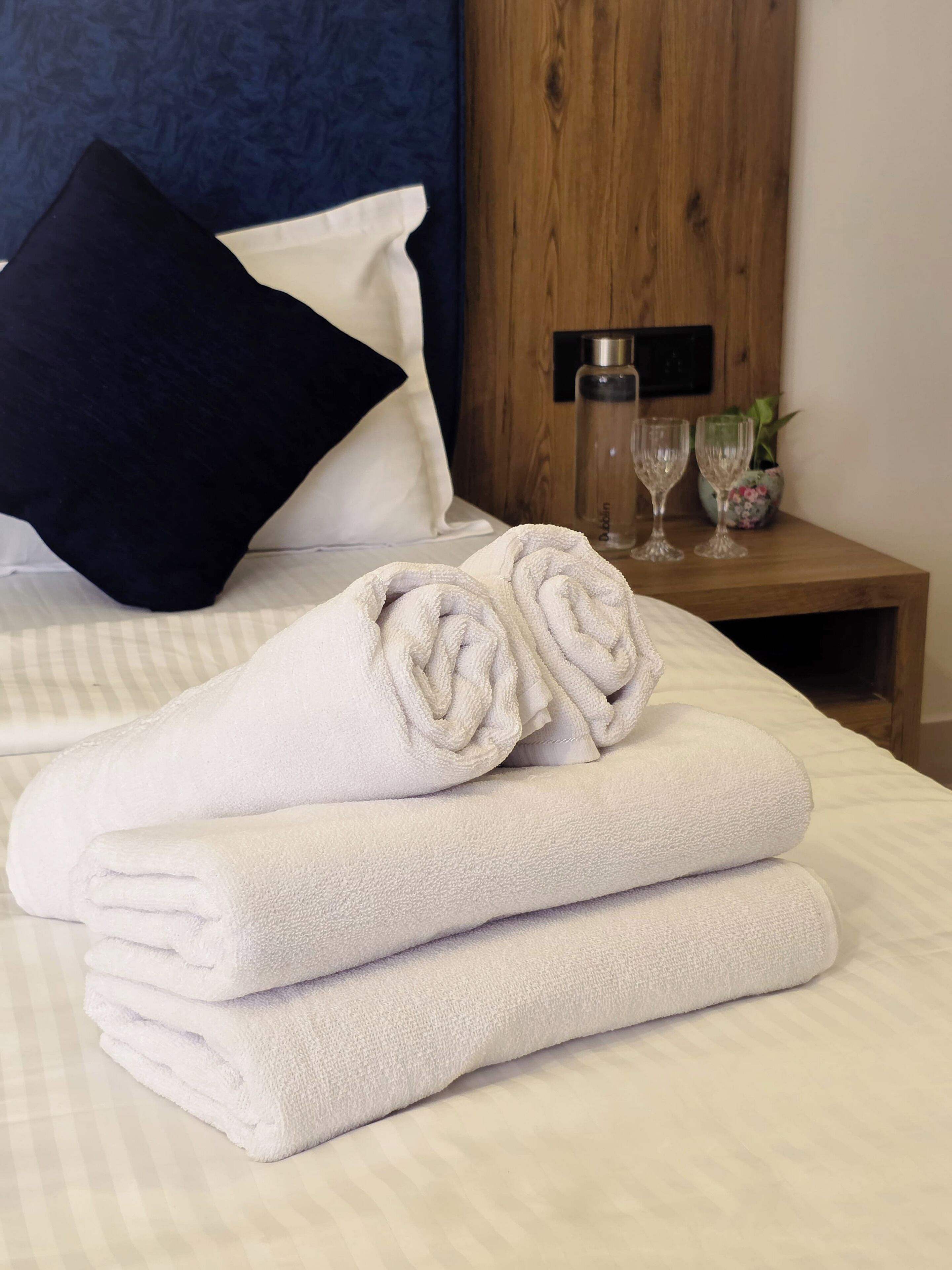 A neatly made bed with folded white towels, a dark blue pillow, and a wooden nightstand with two glasses and a bottle.