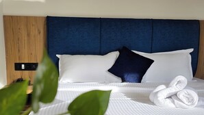 A neatly made bed with white linens and a blue headboard, a rolled towel on the bed, and a potted plant in the foreground. - Ross Harriet View (Port Blair)