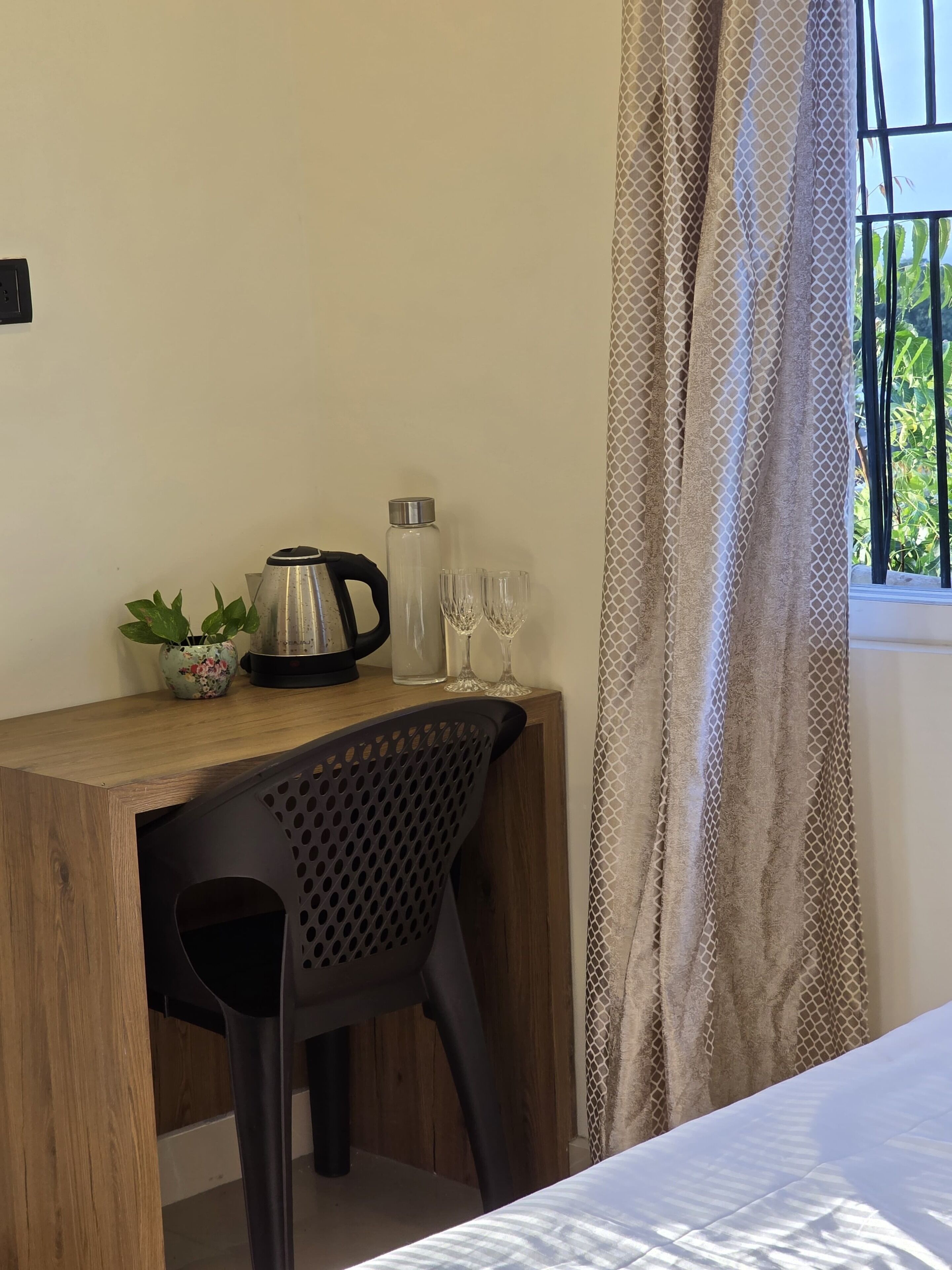 A small, neatly arranged room with a wooden desk, a black chair, a potted plant, a kettle, two glasses, and a bottle.