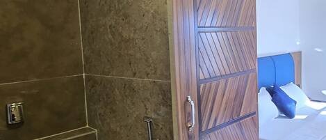 A bathroom with a shower, toilet, and a wooden door leading to a bedroom with a bed and pillows.
