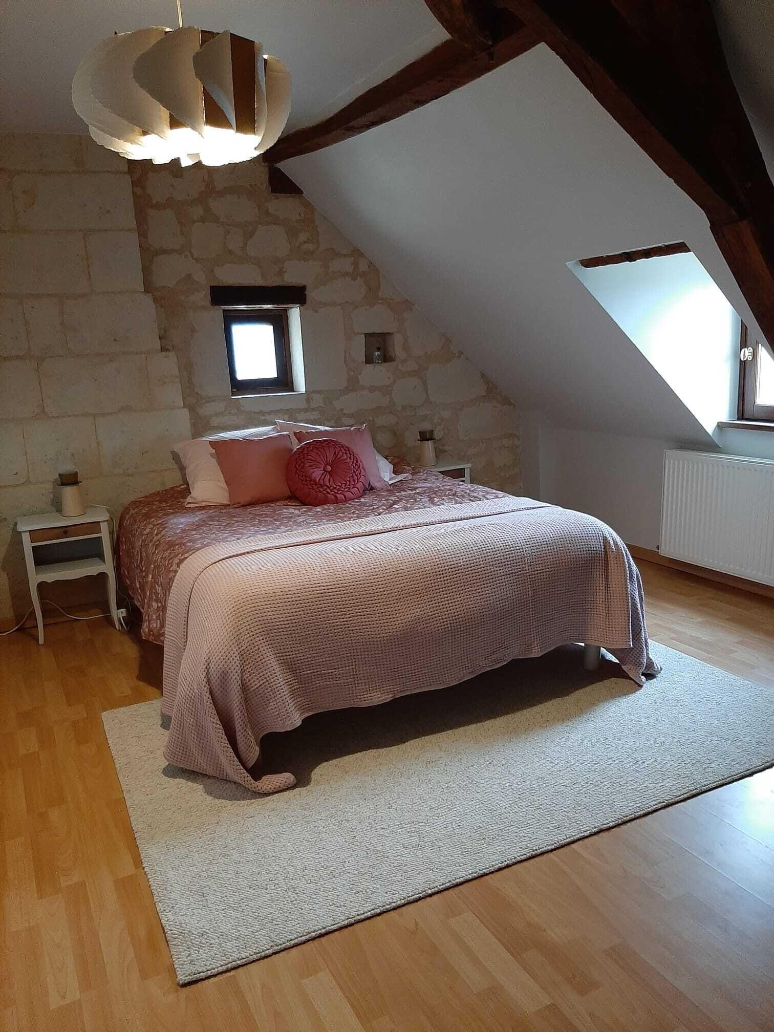 Un dormitorio con techo inclinado, pared de piedra, cama con colcha rosa, mesita de noche, lámpara y ventana con vistas.