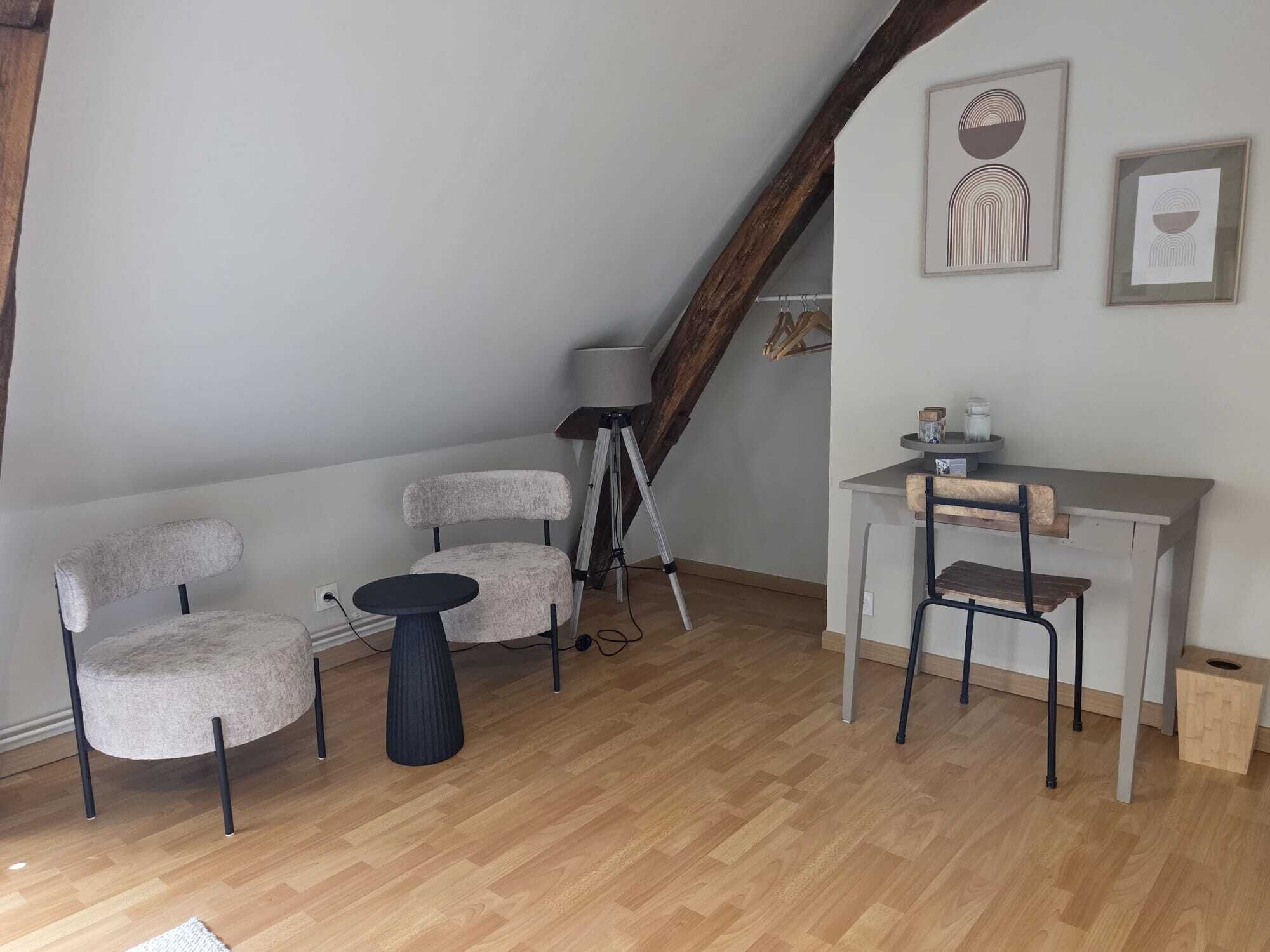 Un dormitorio estilo loft acogedor con techo inclinado, vigas de madera, una pequeña mesa redonda, dos sillas, un escritorio y una lámpara con trípode.
