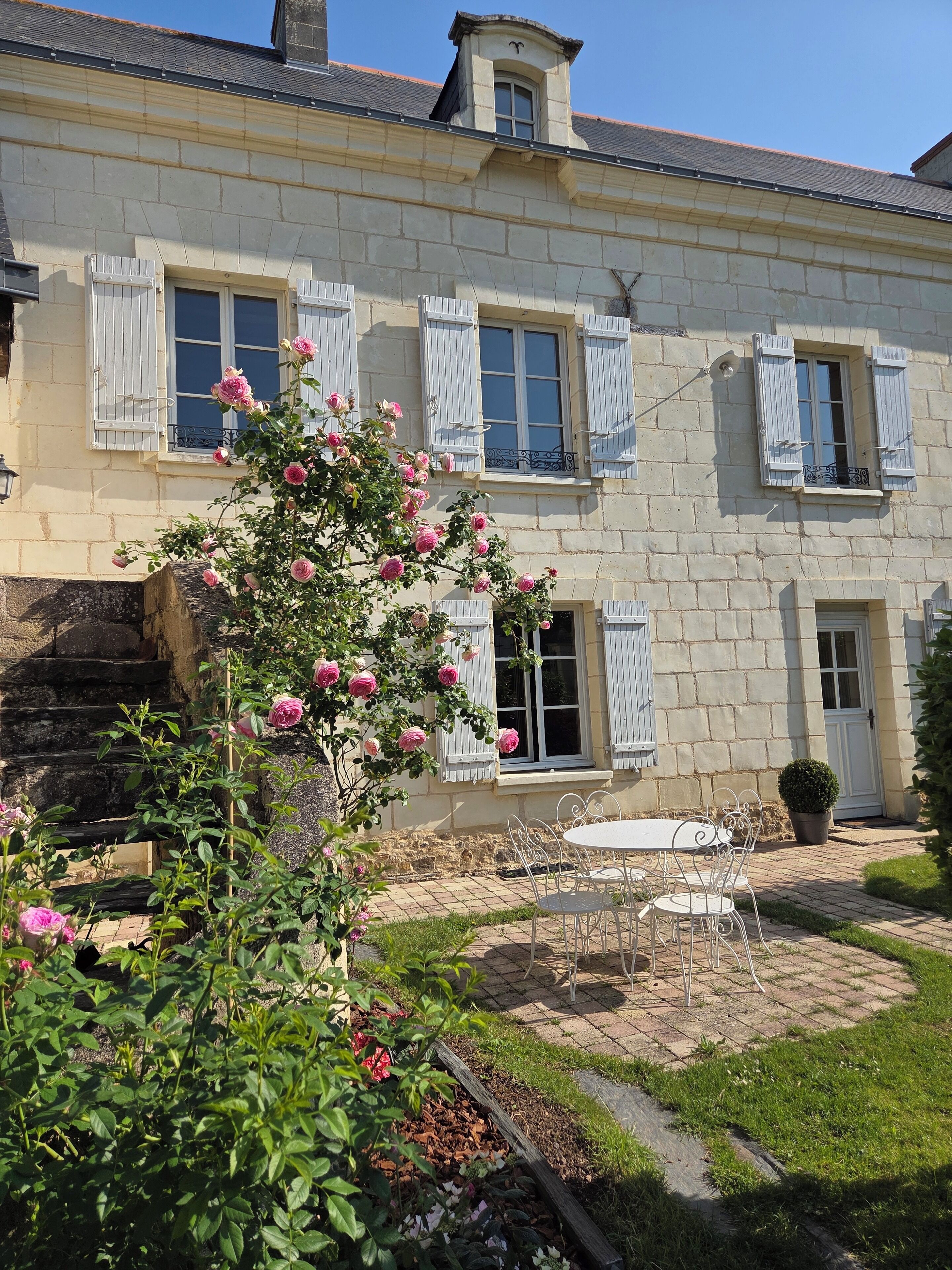 A two-story stone building with white shutters, a garden with roses, and a paved patio with metal chairs.
