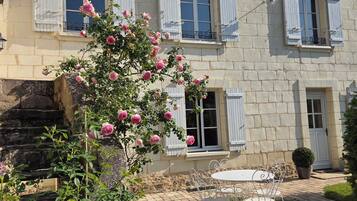 A two-story stone building with white shutters, a garden with roses, and a paved patio with metal chairs.